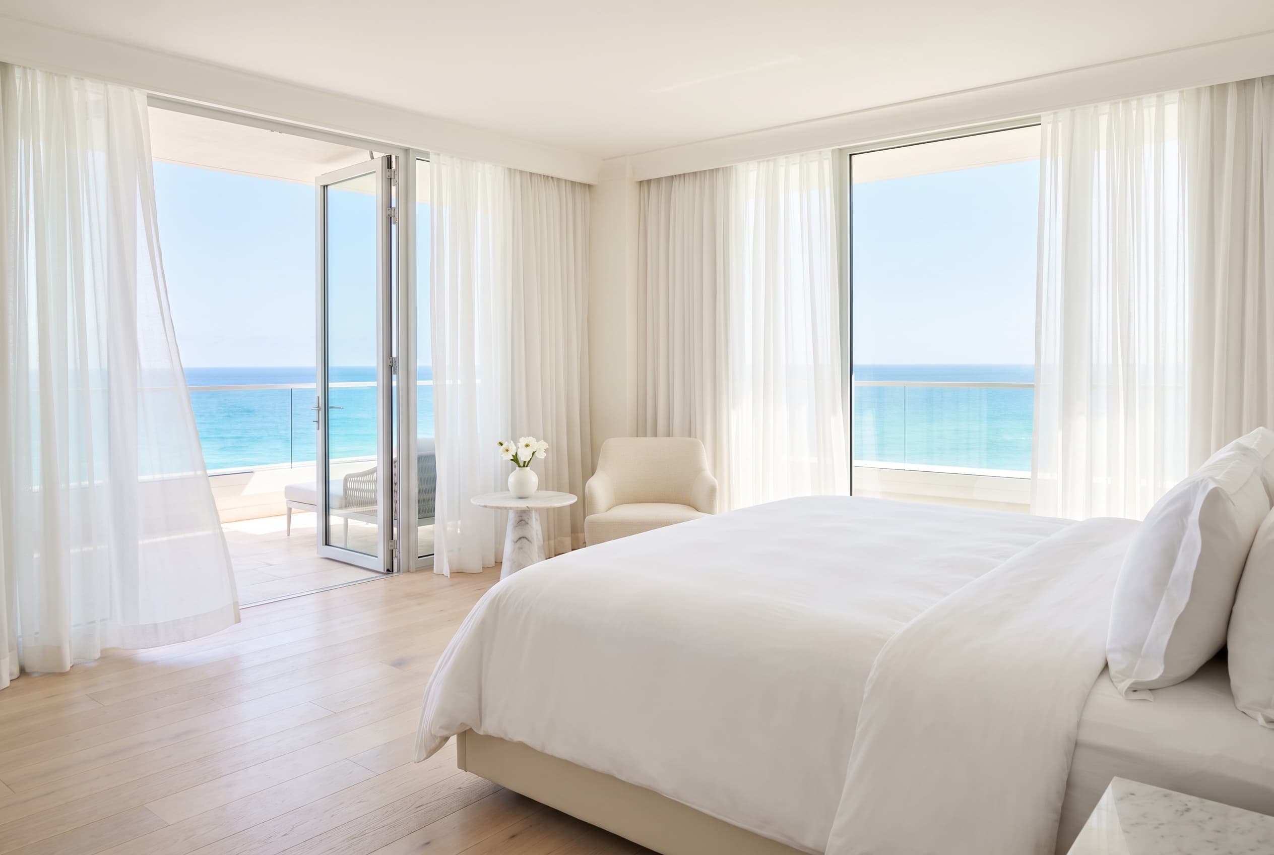 Bright white hotel suite in morning light with balcony doors open to a turquoise sea